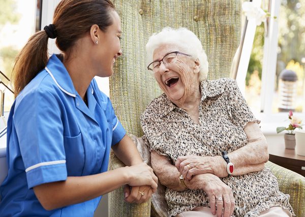 Senior Woman Sitting In Chair And Laughing With Nurse In Retirement Home